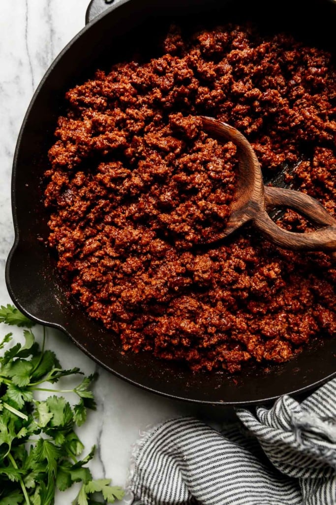Overhead view of cooked ground beef with taco seasoning mixed in, in a cast iron skillet with a twisted handle wooden spoon in the mixture.