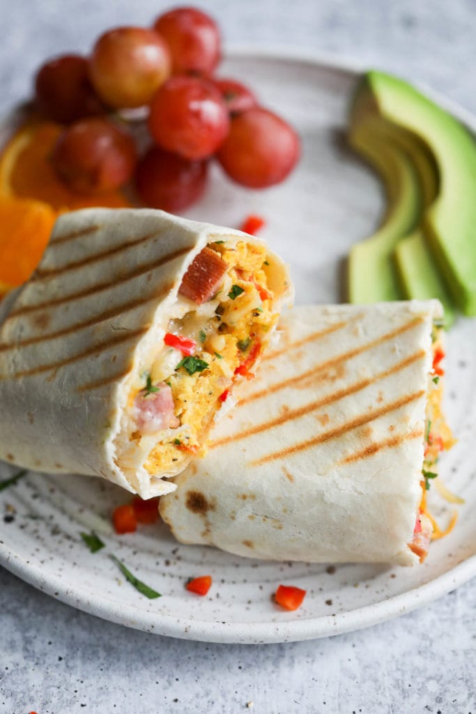 A make-ahead breakfast burrito cut in half showing the scrambled egg, ham, and pepper filling on a speckled plate with fruit served next to it. 