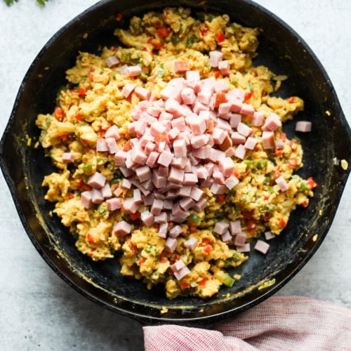 Overhead view of scrambled eggs in a cast iron skillet with diced ham added to the top