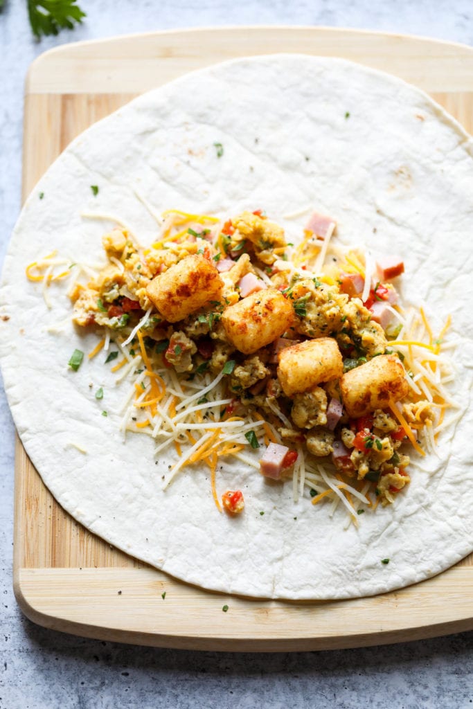 A tortilla laying flat on a wooden cutting board filled with scrambled egg and ham mixture and topped with four tater tots