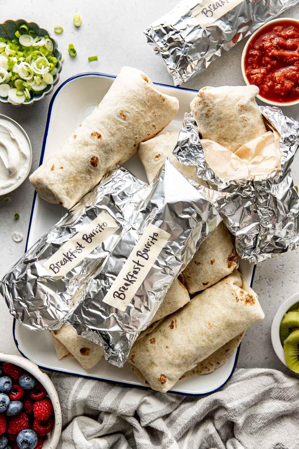 Overhead view of several breakfast burritos on a large platter showing some wrapped in parchment paper and tin foil and labeled 'breakfast burritos"