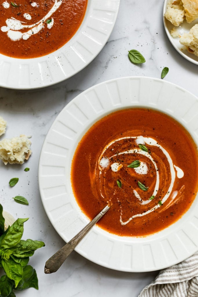 Two white bowls filled with creamy tomato basil soup and topped with a swirl of creme fraiche, black pepper, and fresh basil.