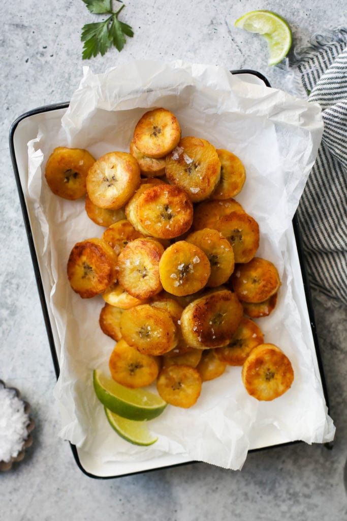 A pile of golden brown fried plantain rounds in a parchment covered dish with sea salt flakes on top.