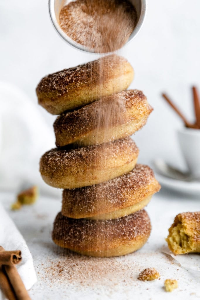 A tall stack of baked donuts with a cinnamon sugar mixture being sprinkled over the top of the stack. 