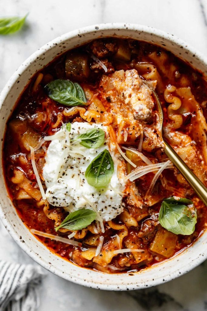 Close up view of creamy lasagna soup with gluten-free ribbon noodles and chunks of ground beef in a bowl topped with ricotta cheese and fresh basil.