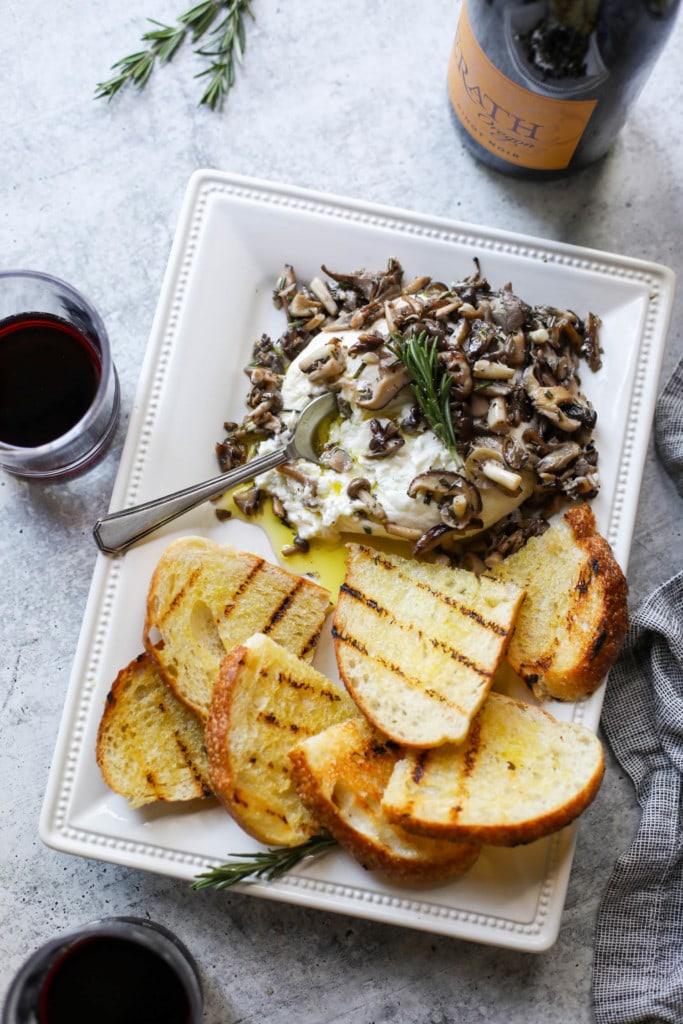 A tray filled with toasted crusty bread, creamy burrata and sauteed mushrooms with a bottle of wine in the background.