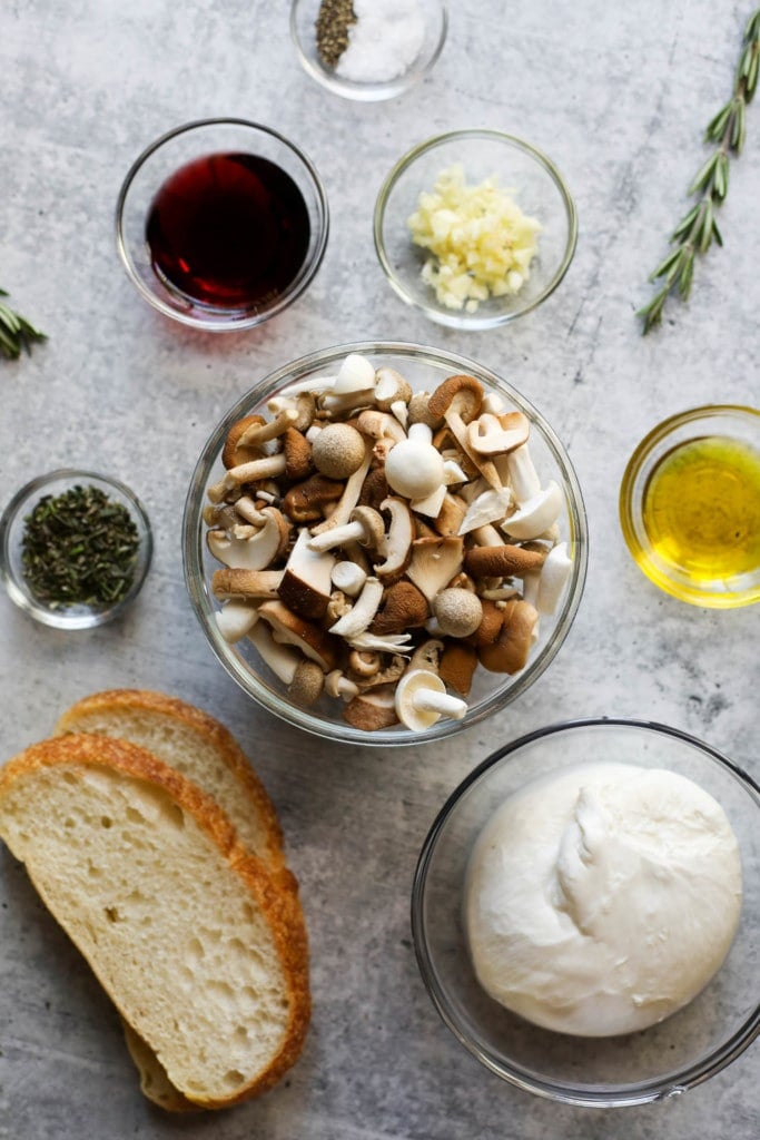 All ingredients for creamy burrata topped toast with mushrooms in bowls along with a short glass of pinot noir.
