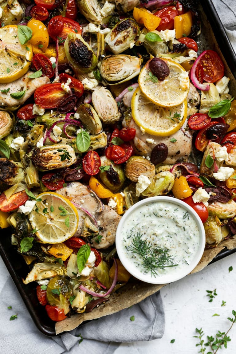 Overhead view Greek chicken thighs with veggies on sheet pan with side of tzatziki sauce.