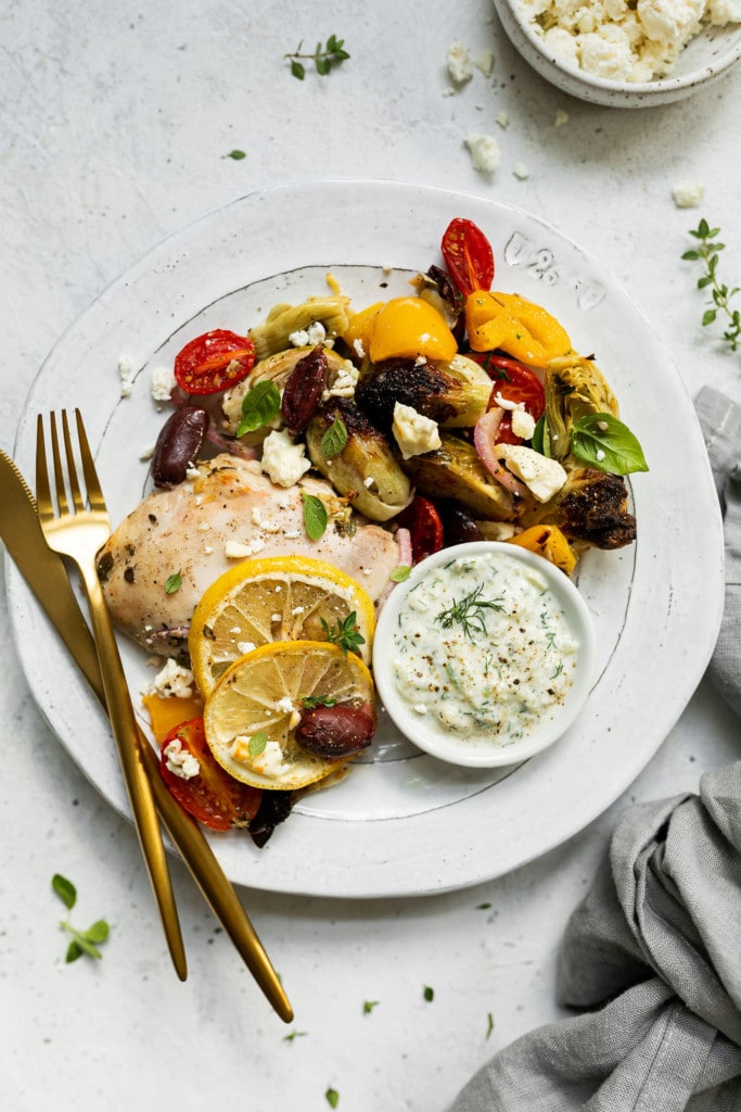 A plate full of roasted Greek chicken and vegetables topped with feta cheese and herbs with a side of tzatziki sauce.