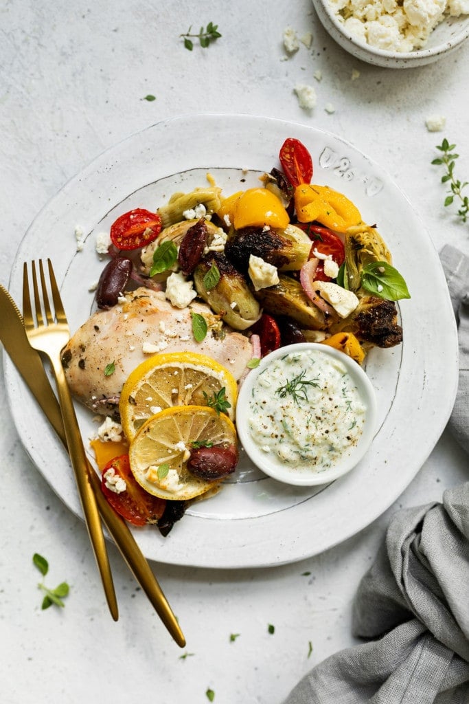 A plate full of roasted Greek chicken and vegetables topped with feta cheese and herbs with a side of tzatziki sauce.