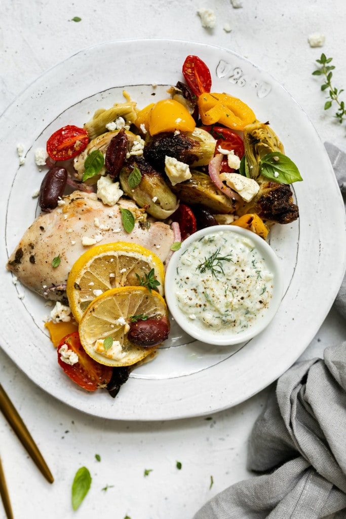 A roasted Greek flavored chicken thigh with a pile of roasted vegetables plated on a white plate topped with feta crumbles and a side of Tzatziki sauce.