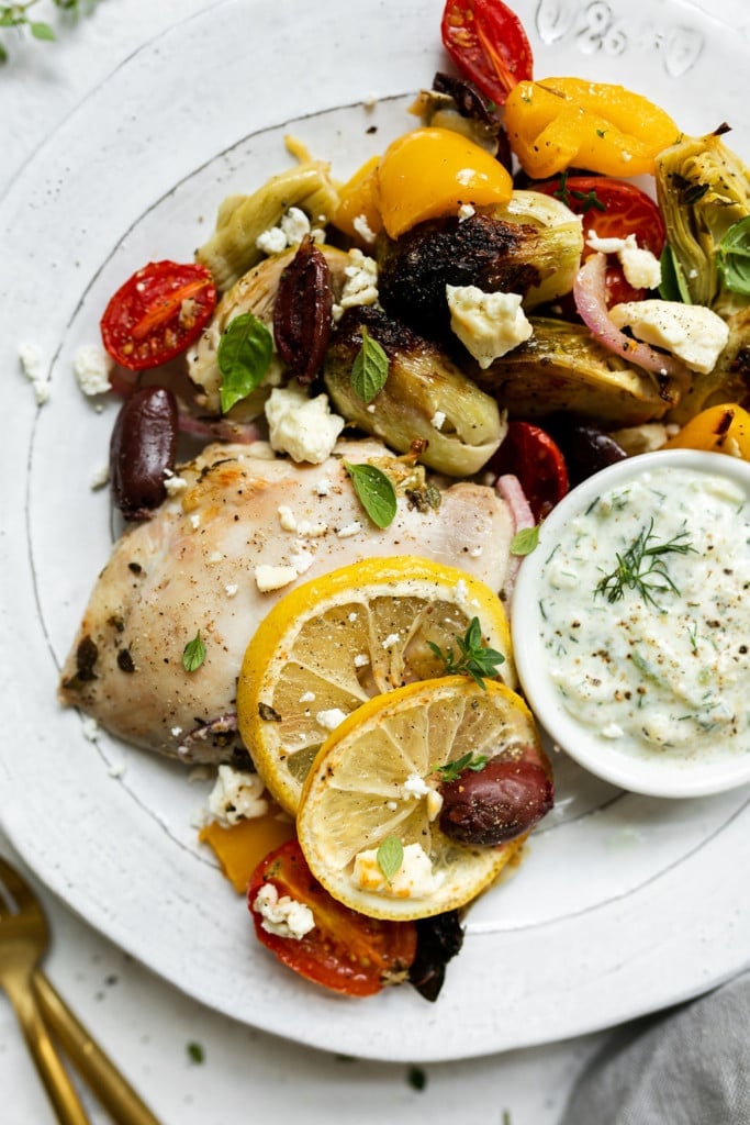 A close up view of roasted Greek veggies, artichoke, tomatoes, kalamata olives, on a plate with a roasted chicken thigh and a side of Tzatziki sauce.