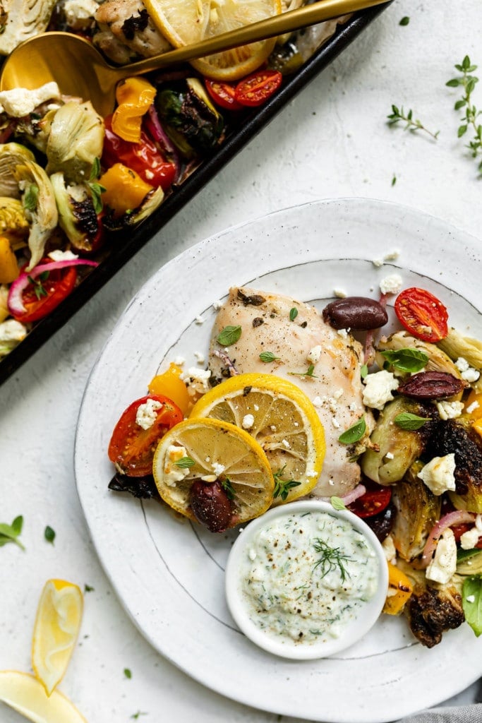 Greek chicken with vegetables plated on a white plate with a side of tzatziki sauce.