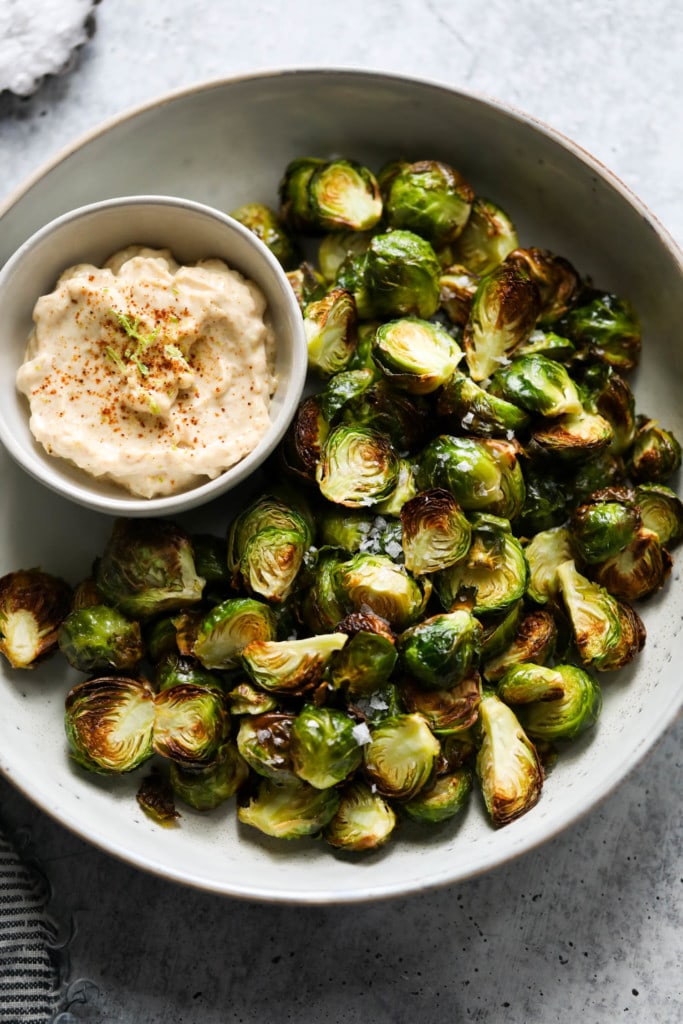 Crispy Brussels sprout halves made in the air fryer plated on a serving tray and sprinkled with sea salt flakes with a bowl of aioli on the side.