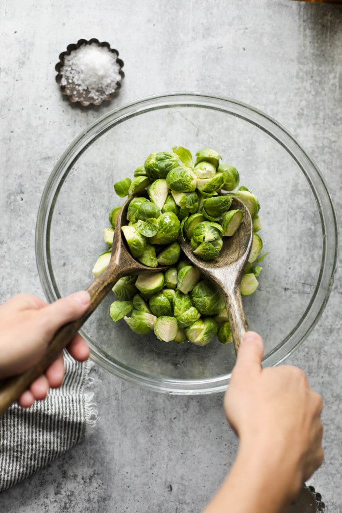 Fresh cut Brussels sprout halves in a clear mixing bowl being tossed with two wooden spoons.