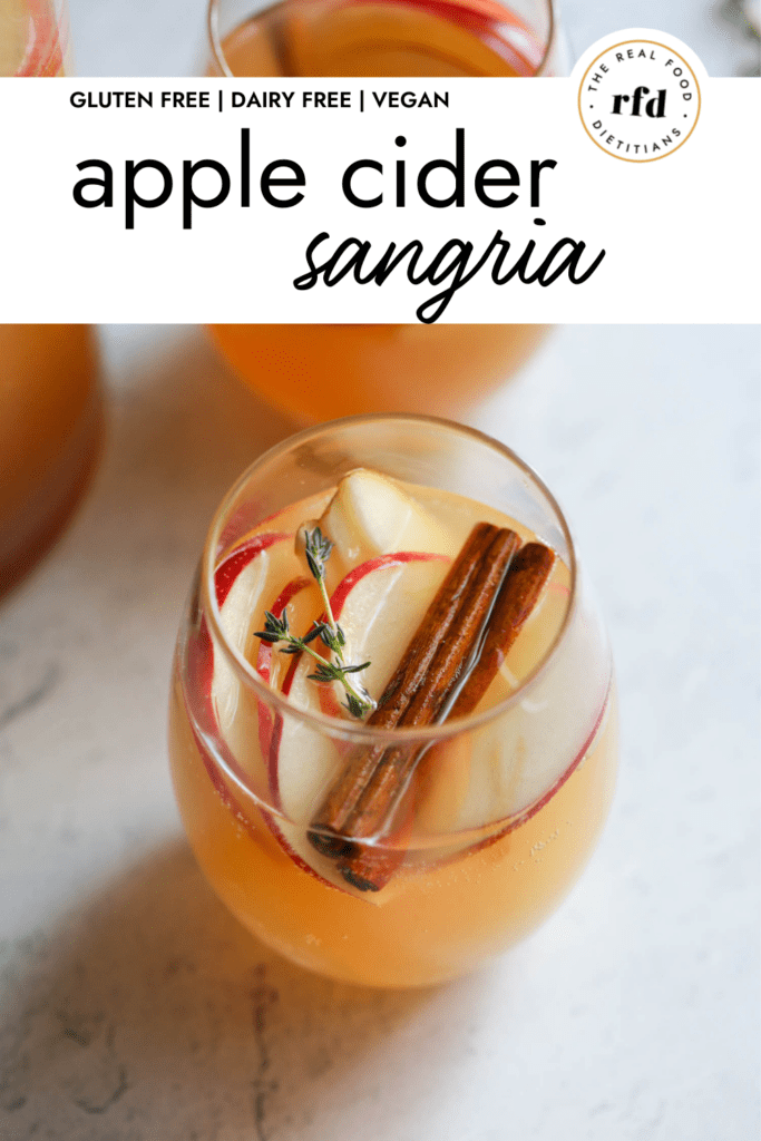 Close up view of a glass of apple cider sangria with apple slices, cinnamon sticks, and fresh thyme floating in the sangria.