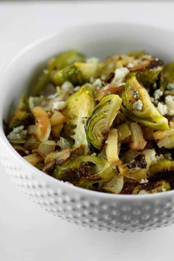 Roasted Brussels sprouts in a white bowl topped with sauteed apples and fresh blue cheese.