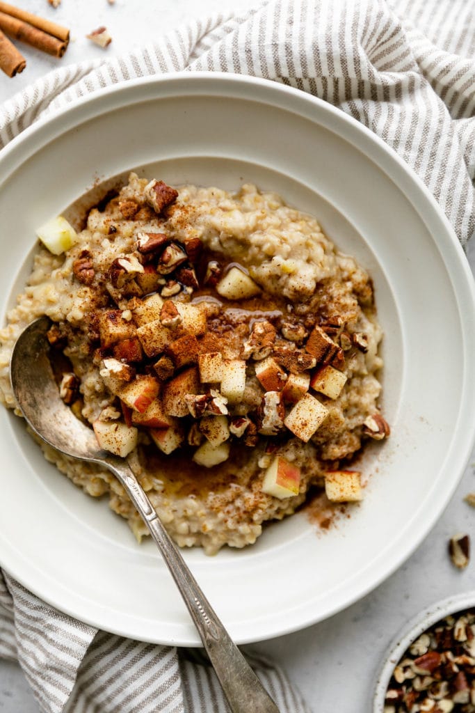 Creamy steel cut oatmeal in a white bowl topped with apple chunks, cinnamon, and pecans. 