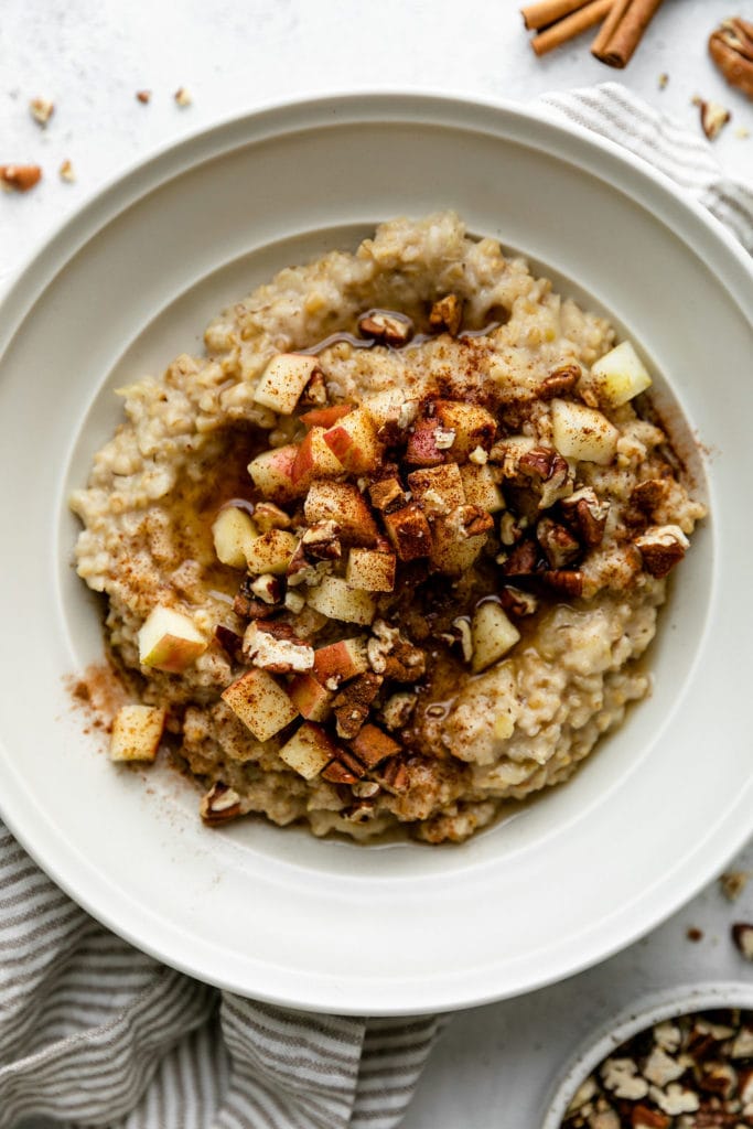 Close up view of creamy steel cut oatmeal in a white bowl topped with apples, pecans, and cinnamon.