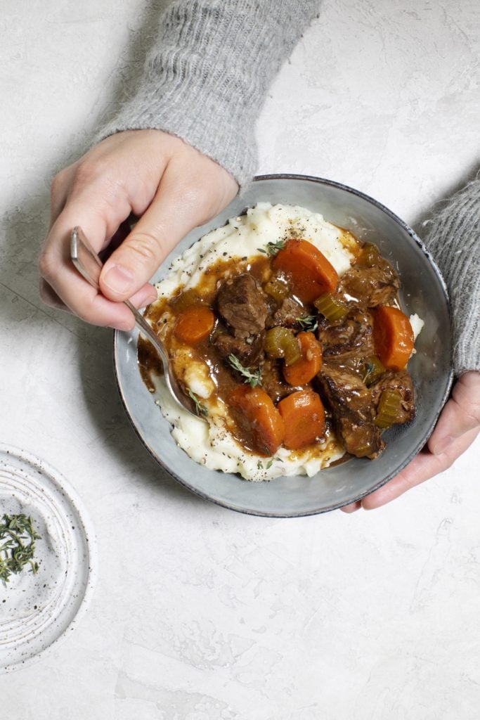 Hands holding a bowl of Irish Beef Stew served over mashed potatoes.