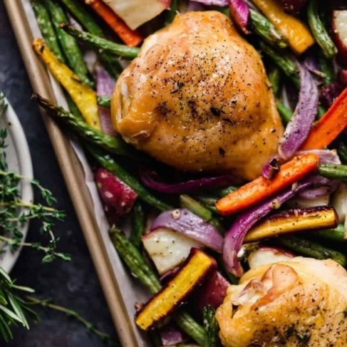 Close up view of a roasted chicken thigh on a plate with colorful roasted vegetables.