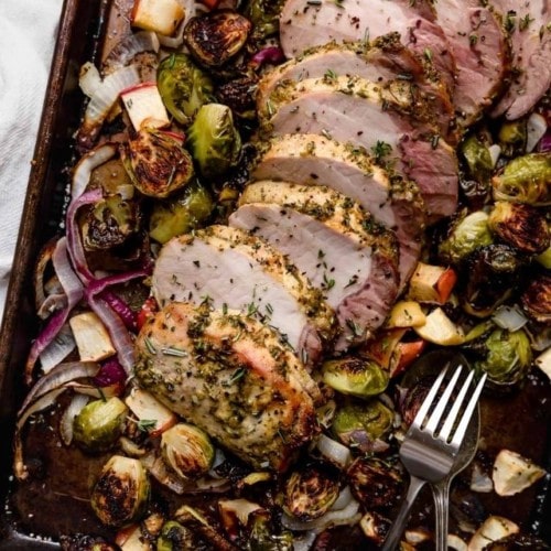 Thick slices of herb-dijon crusted pork loin on a baking sheet with Brussels sprouts and apples.