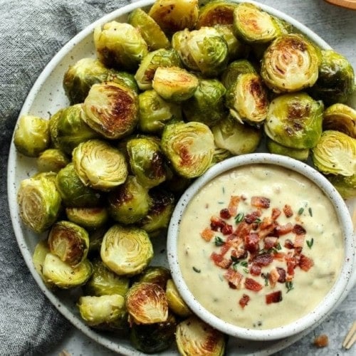 A platter of roasted Brussels sprout halves with a small bowl of garlic aioli topped with bacon crumbles