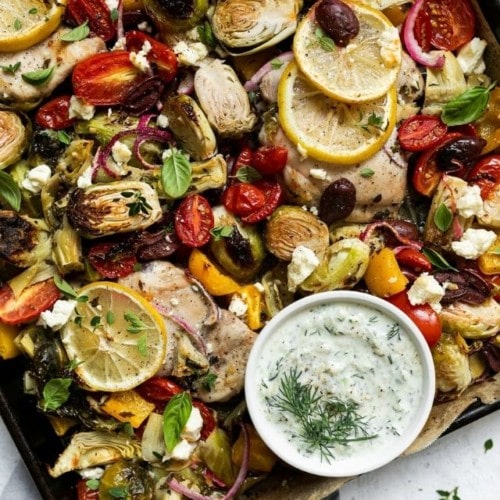 A sheet pan filled with chicken thighs, artichokes, tomatoes, and Brussels sprouts topped with Greek seasonings.