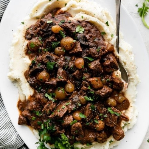 Fork tender beef tips in a saucy gravy with pearl onions and mushrooms served over a large platter of mashed potatoes.