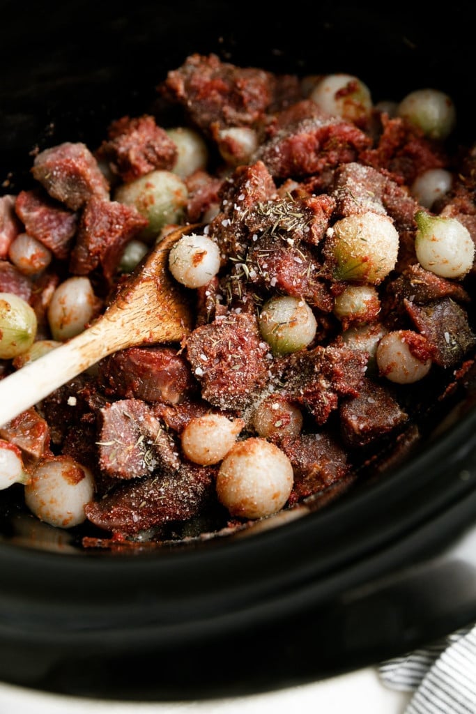 A crockpot filled with beef tips, pearl onions, and sauce ready to begin slow cooking.