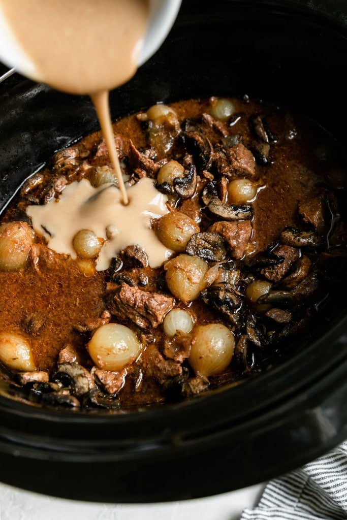 Overhead view of beef tips cooked down in a slow cooker with a bowl of gravy being poured into the beef.