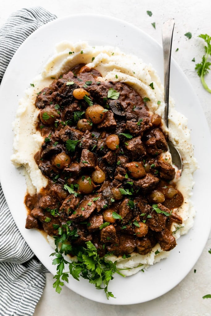 Slow cooker beef tips with gravy served over a large platter of creamy mashed potatoes.