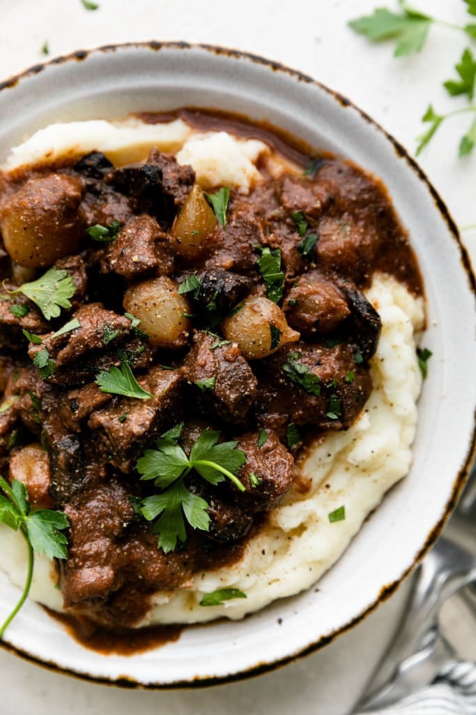 Slow cooker beef tips with pearl onions in homemade gravy served over a high pile of creamy mashed potatoes and garnished with fresh herbs.