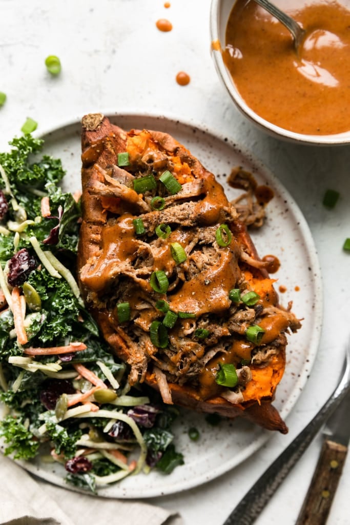 Shredded pork in a baked sweet potato topped with honey mustard sauce and green onions plated with a kale side salad.