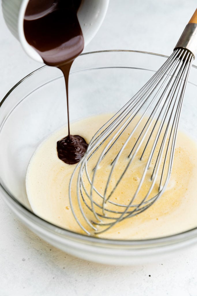 Melted chocolate being poured into the peppermint brownie batter in a glass bowl with a whisk.