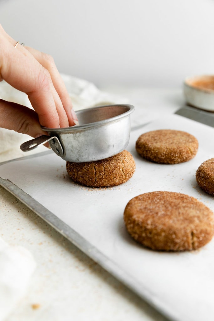 Snickerdoodle cookies lined up on a parchment-covered baking sheet being flattened with a silver measuring cup