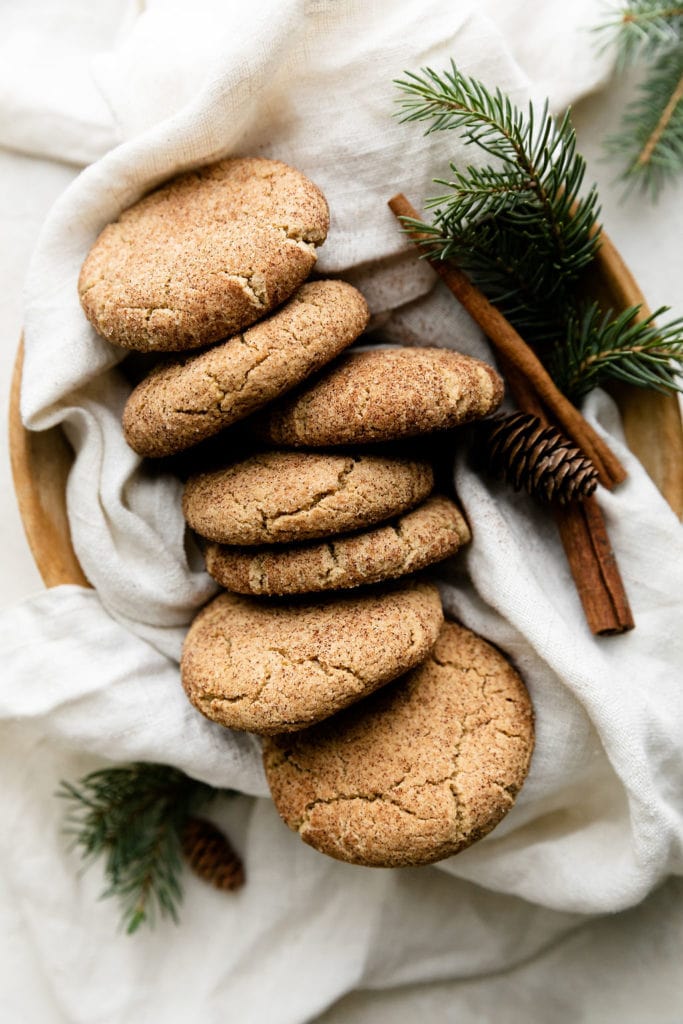 Several Snickerdoodle cookies with cinnamon sugar coating lined up in a wooden tray with a white cloth, cinnamon stick, pinecone, and greenery.