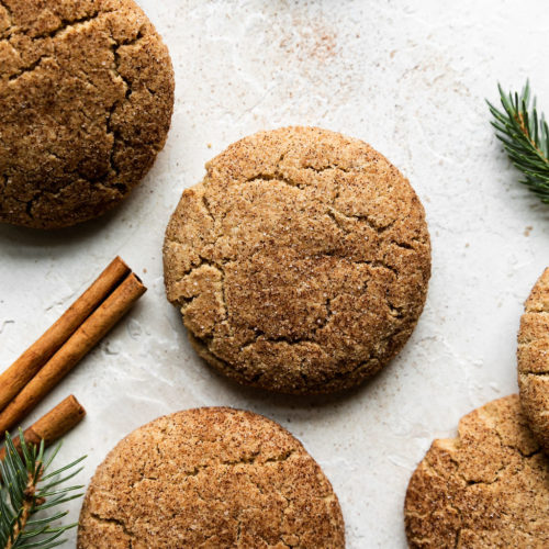 Several gluten-free snickerdoodle cookies sprinkled with cinnamon sugar on a counter with sprigs of greenery nearby.