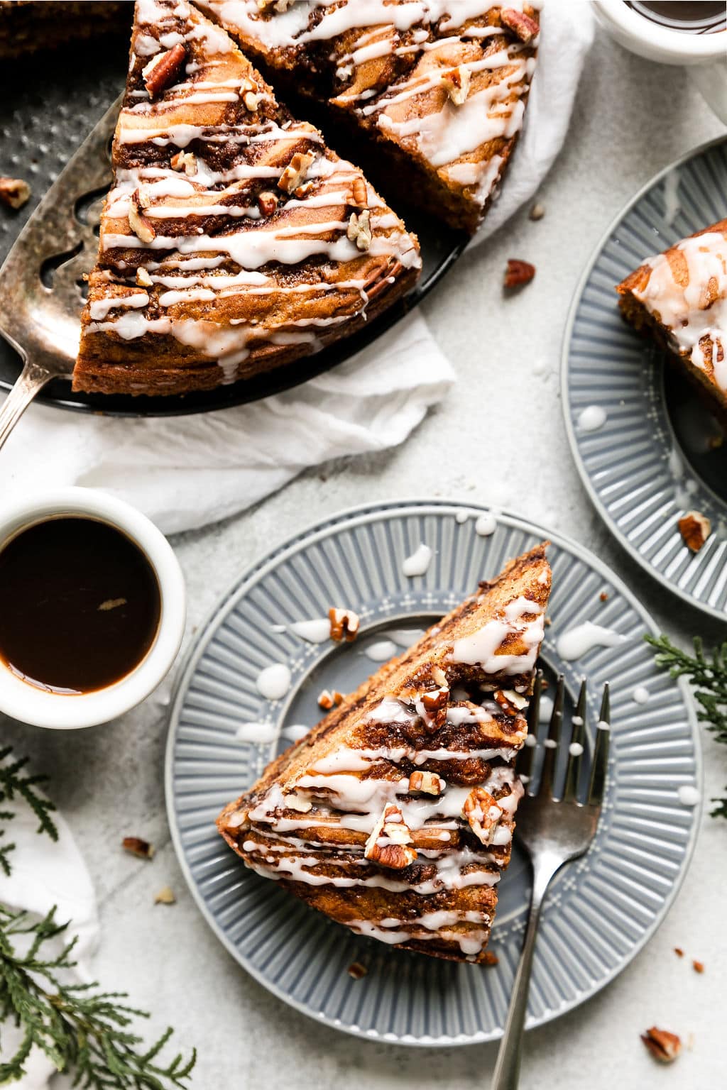 A slice of cinnamon roll coffee cake with white glaze and chopped pecans on top being lifted from a round cake with server, slices on blue plates. 