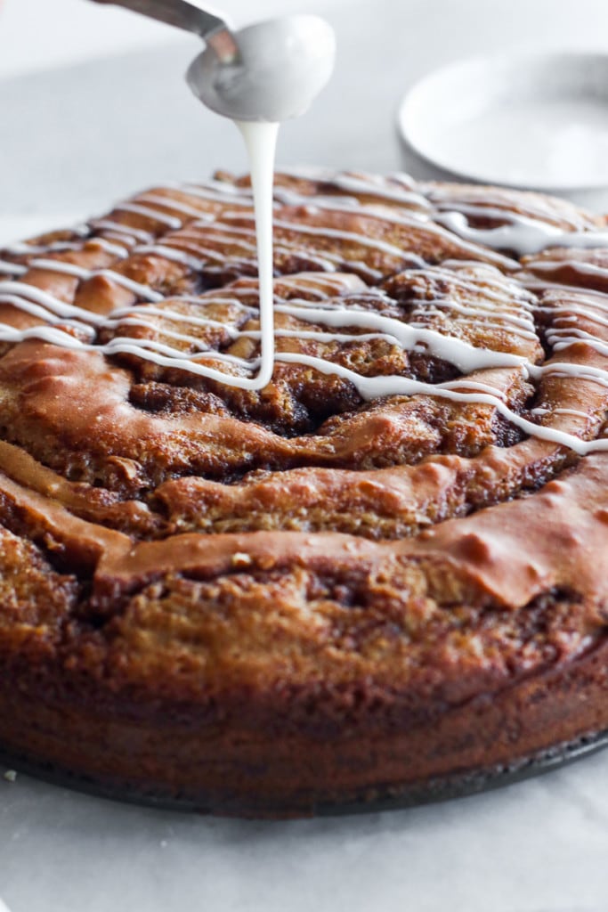 A white frosting glaze being drizzled over a round gluten free cinnamon roll coffee cake.