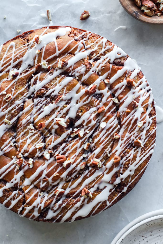 Overhead view of a round cinnamon roll coffee cake with a white frosting drizzle and chopped pecans sprinkled on top.