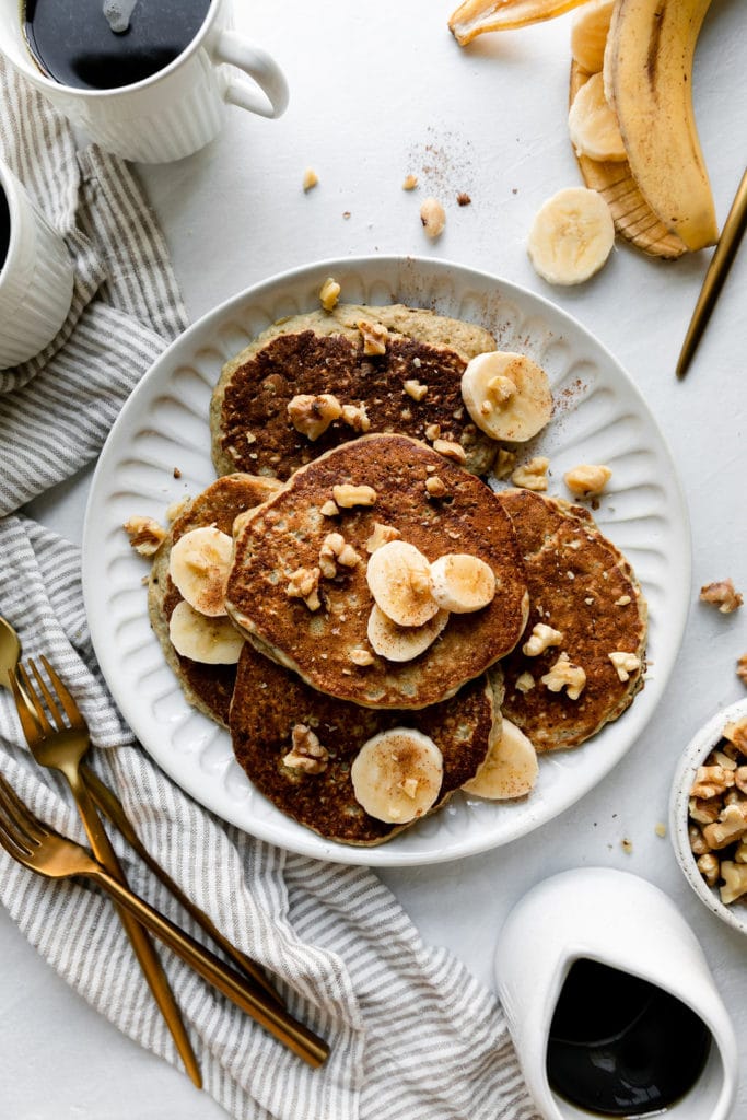 Overhead view of a white plate filled with several banana oatmeal pancakes topped with banana slices, walnuts, and cinnamon.