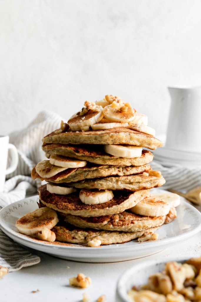 A tall stack of banana oatmeal pancakes on a white plate with banana slices between the layers of pancakes.