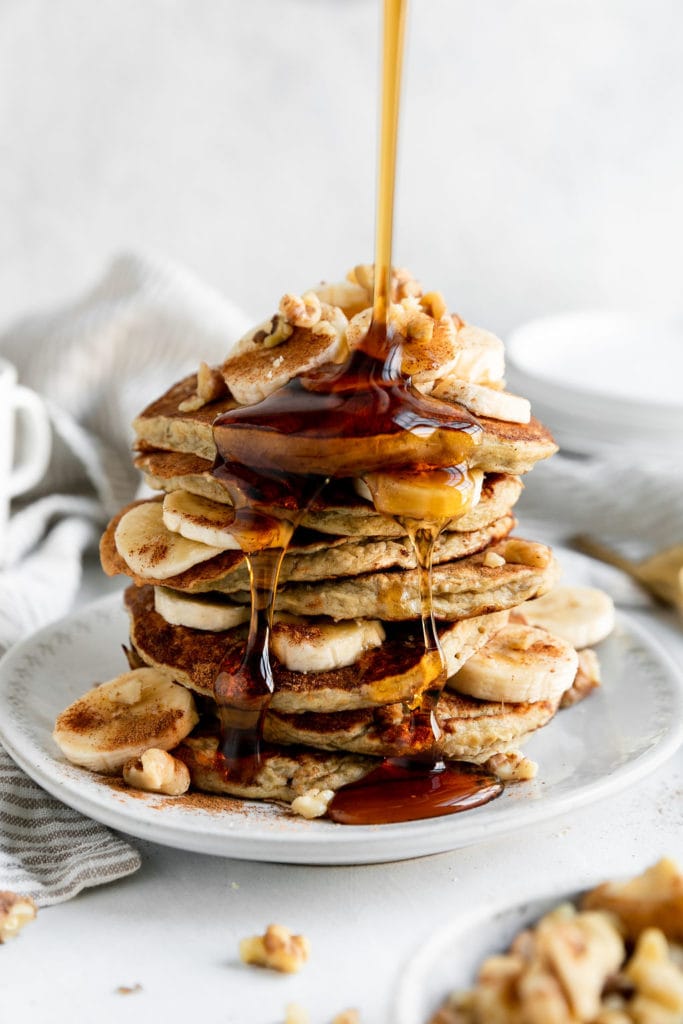 A tall stack of banana oatmeal pancakes with banana slices between the stacks, thick maple syrup being poured over the stack and running down the side.