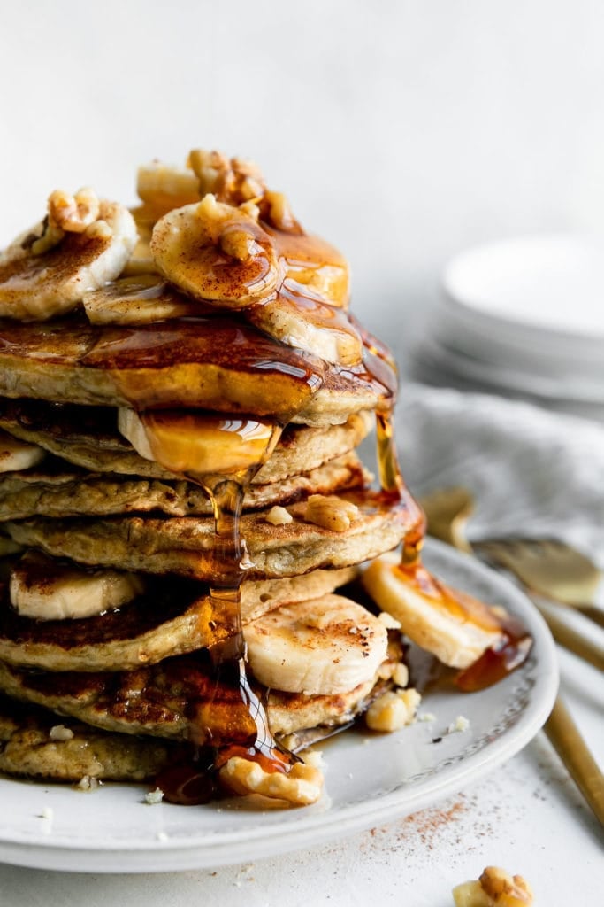 Close up view of a banana oatmeal pancakes stacked tall on a plate, topped with banana slices, walnuts, cinnamon, and thick maple syrup.