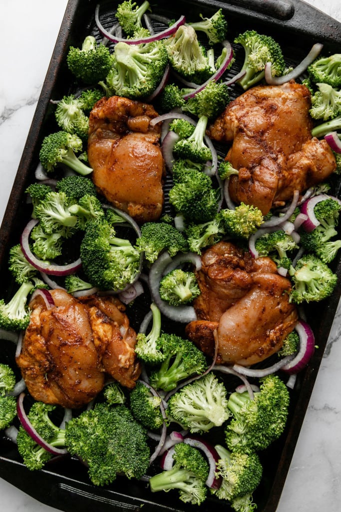 Chipotle chicken thighs with broccoli and red onion arranged on a baking sheet ready for the oven.