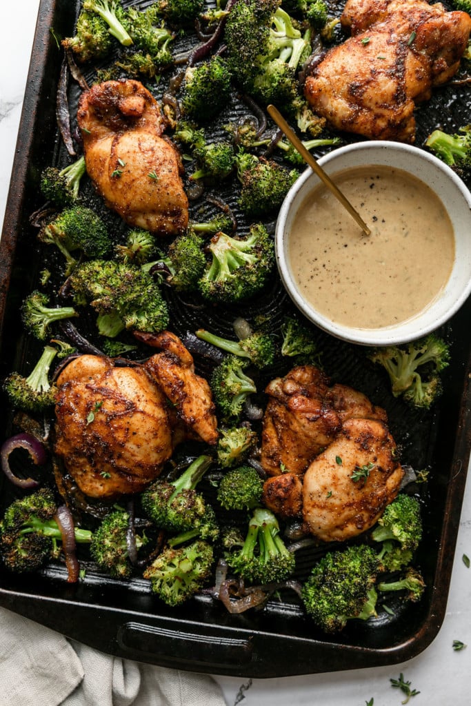 Overhead view of freshly baked chipotle chicken thighs on a sheet pan amongst crispy baked broccoli with a small bowl of homemade honey mustard sauce on the side. 