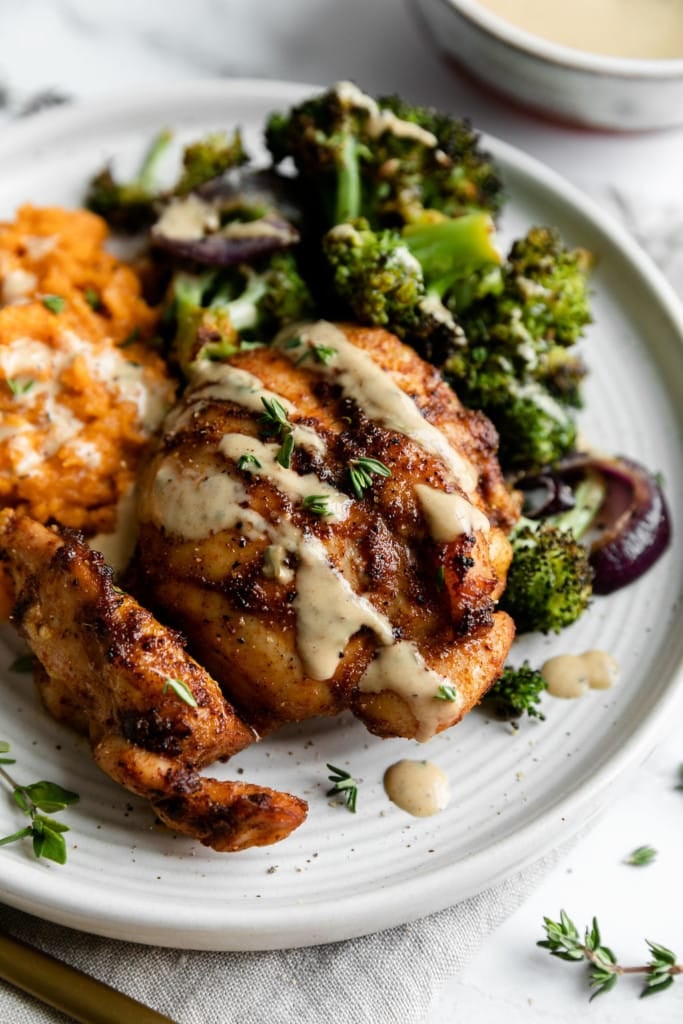 Close up view of chipotle chicken thigh drizzle with honey-mustard sauce and fresh herbs plated on a white plate with a side of broccoli and mashed sweet potatoes. 
