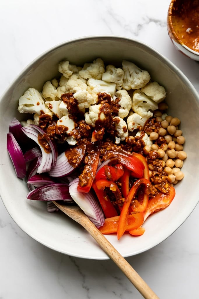 Cauliflower, chickpeas, red onion, and orange peppers in a bowl with spices sprinkled on top.