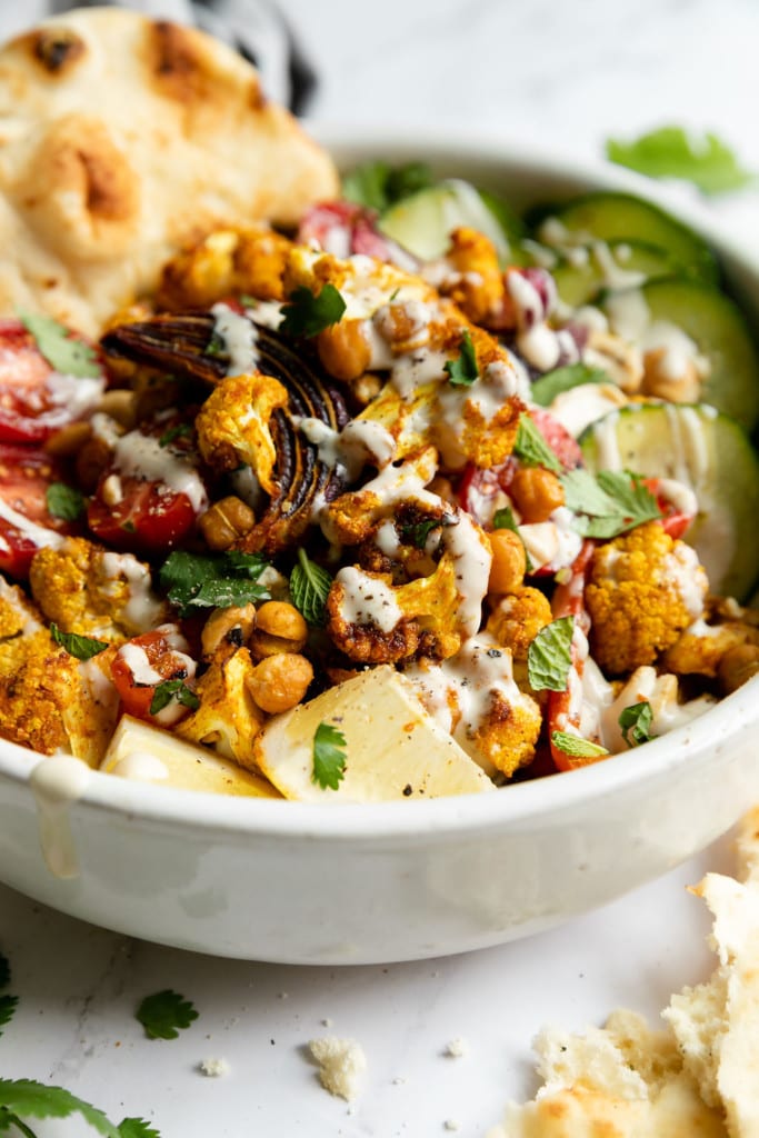 Close up view of cauliflower and chickpea shawarma in a bowl with mixed greens, cucumbers, and lemons with tahini dressing drizzled on top.